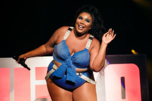 Lizzo waving on stage in a blue denim bustier and shorts, holding a microphone