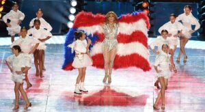Jennifer Lopez on stage in a silver outfit, a feathered Puerto Rican flag cape behind her and children in white around her