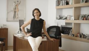 Lourdes López in a black top and white trousers, leaning on a desk in her office