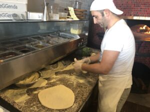 A pizza maker in a white T-shirt stretching dough beside a wood-fired oven