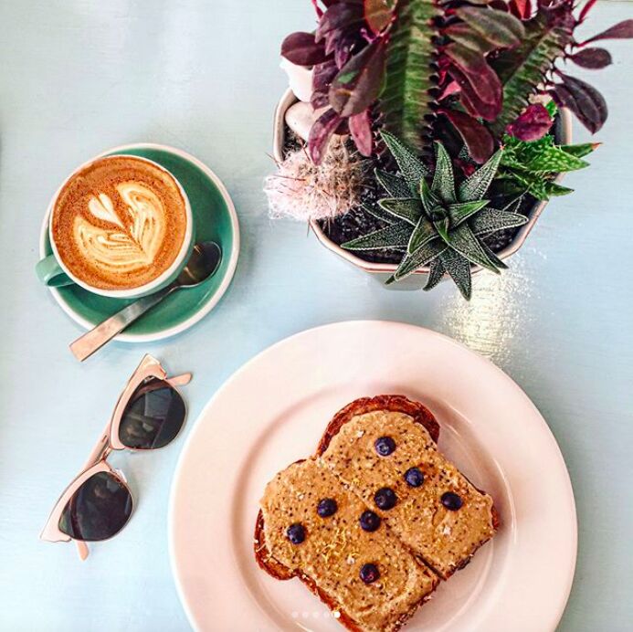 A latte with leaf art, toast with nut butter and blueberries, sunglasses and a potted succulent on a pale blue table