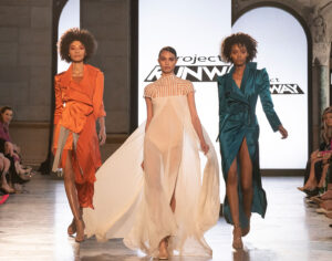 Three models walking a Project Runway catwalk in orange, cream and teal outfits