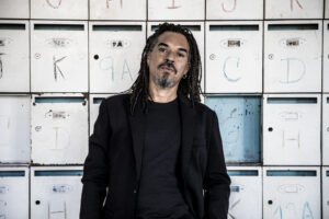 X Alfonso in black with long dreadlocks, standing in front of a wall of lettered mailboxes
