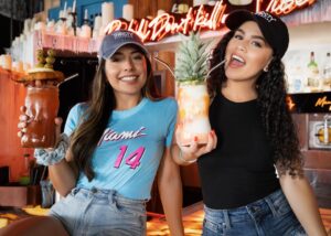 Two women laughing in a bar, holding up drinks in a tiki mug and a pineapple
