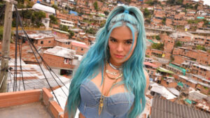 Karol G with long turquoise hair and a denim corset, on a rooftop above a hillside neighbourhood