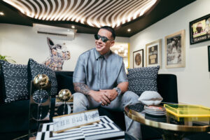 Daddy Yankee in sunglasses and a grey suit, sitting on a sofa in a room with a striped ceiling and gold records on the wall