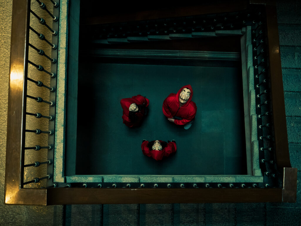 Looking down a stairwell at three people in red jumpsuits and Dalí masks