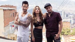 The stars of La Reina del Flow, two men and a woman posing on a rooftop above a hillside city