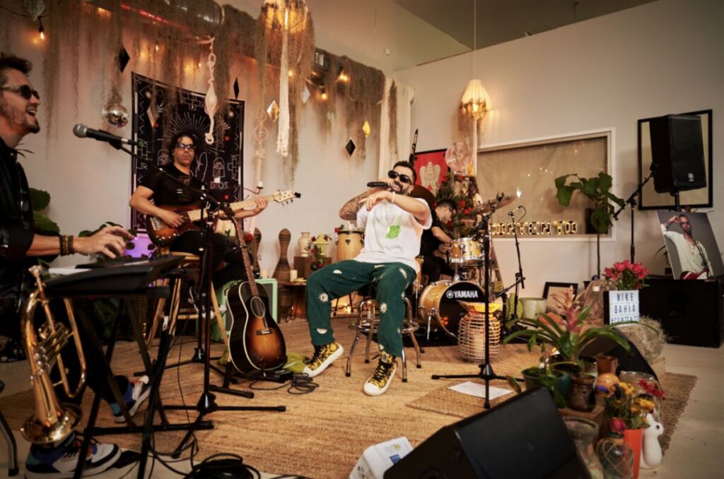 Mike Bahía playing with a band in a room decorated with hanging lights and plants