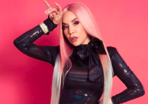 Ivy Queen with long pink hair and a black patent-leather top, pointing to her head, against a pink wall