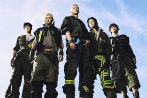 NCT 127, six members in utility outfits and combat boots, photographed from below against a blue sky