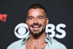 Ricky Martin smiling in an open-collared shirt and necklaces in front of a CBS logo