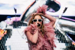 Suki Waterhouse performing in a pink ruffled dress and white sunglasses, hands in her hair