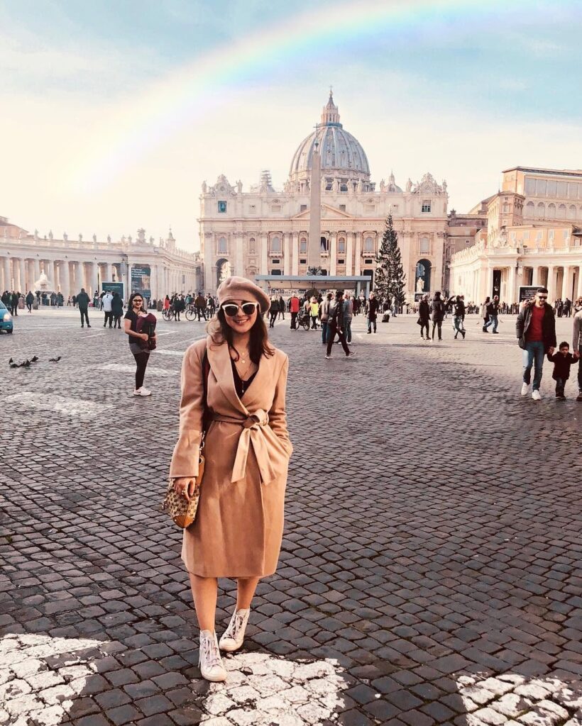 Cata in a camel coat and beret on the cobbles of St Peter’s Square, the basilica’s dome behind her and a rainbow arcing across the sky.