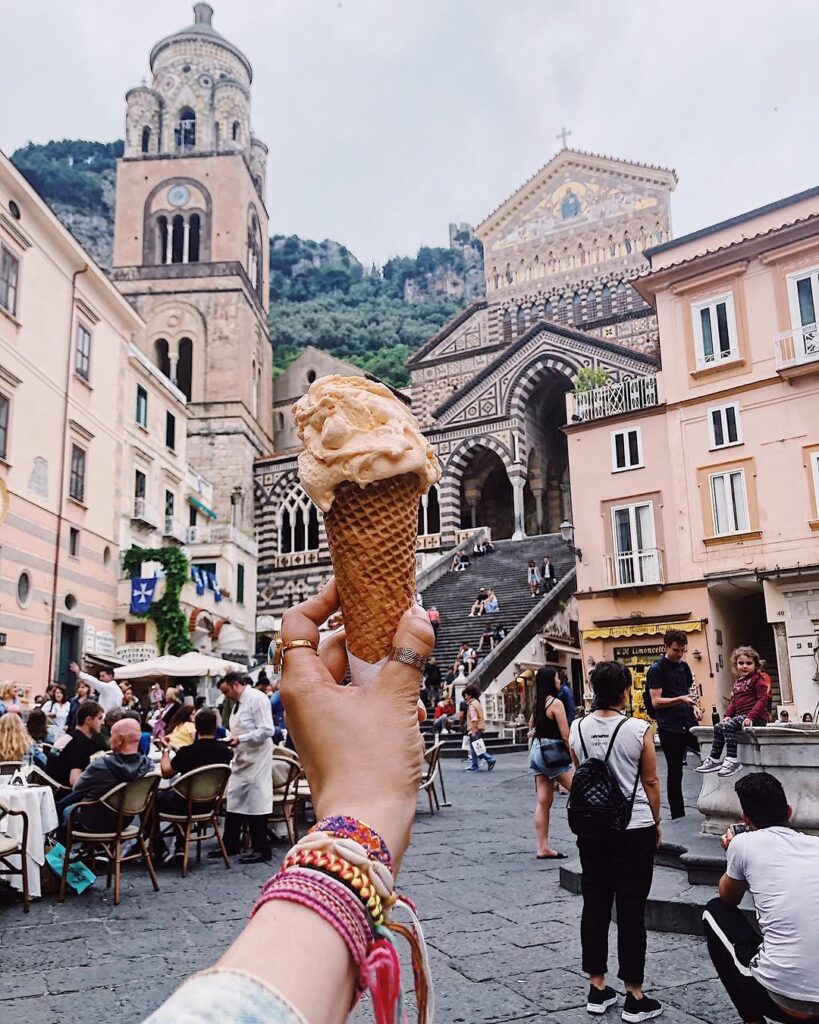 A hand holding a gelato cone up in front of Amalfi’s cathedral, its striped stonework and wide staircase behind, the piazza busy below.