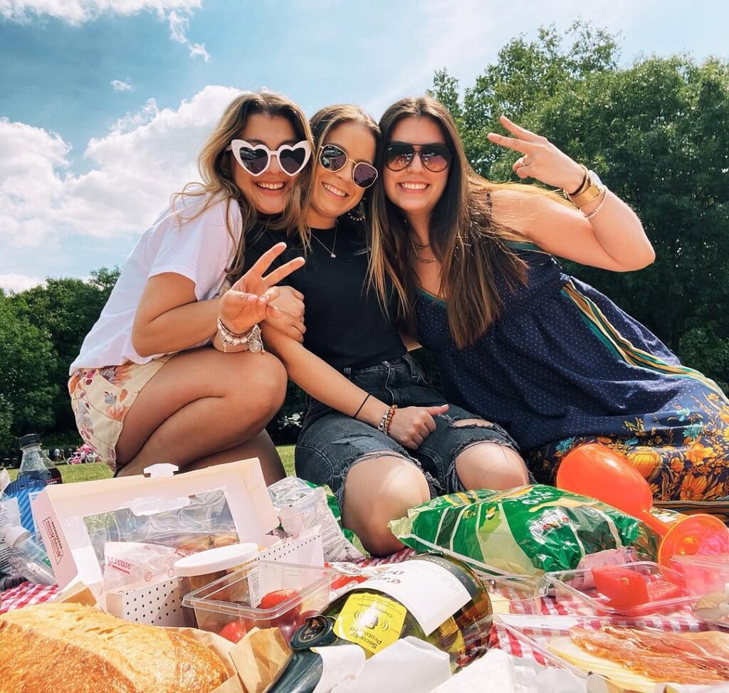 Three friends kneeling on a picnic rug in a London park throwing peace signs in sunglasses, bread, cheese, tomatoes and wine spread in front of them.