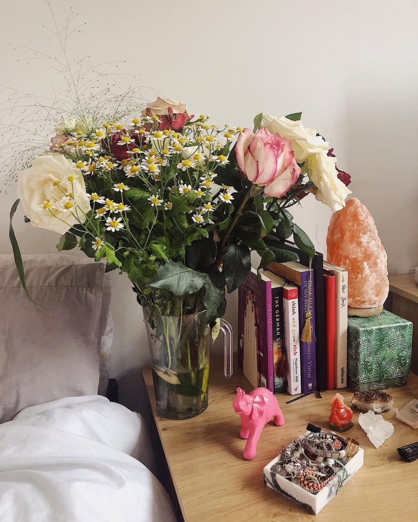 A bedside table beside white linen: a glass jar of roses and daisies, a row of paperbacks, a salt lamp, crystals and a small pink elephant.