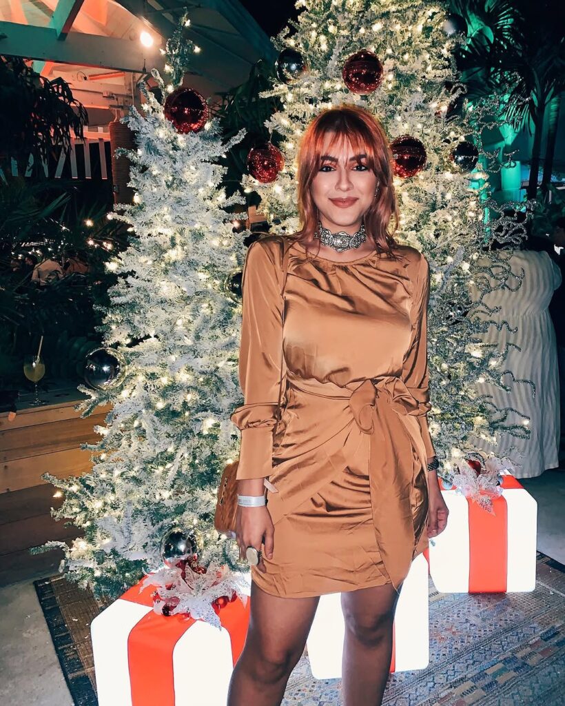 Cata in a caramel satin mini dress with a knotted waist, in front of a lit Christmas tree hung with red baubles, oversized gift boxes at her feet.