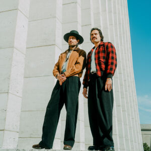 Hermanos Gutiérrez, brothers Alejandro and Estevan Gutiérrez, standing in front of tall white stone columns under a blue sky.